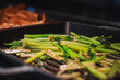 © Aerial Film Studio - Close up of glossy green stems sizzling on a ridged grill pan in Japan. Light catches char and steam, with browned root slices softly blurred in the background.