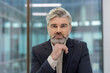 © Liubomir - Mature businessman confidently posing in an office, resting his chin on his hand while looking directly creating a portrait of professionalism, experience, and executive leadership
