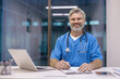© Liubomir - Smiling male doctor wearing blue scrubs and stethoscope, sitting at a desk while writing on a clipboard, representing professionalism and dedicated healthcare services