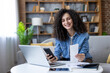 © Liubomir - Smiling woman holding a paper bill, using a smartphone and laptop to manage her personal budget and financial planning from a comfortable home setting