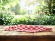 © Milana - Red and white checkered cloth folded on a wooden table with lush green garden background and soft sunlight creating a peaceful outdoor atmosphere