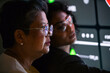 © artitwpd - Two professionals collaborate beside a glowing digital dashboard, reflections on their glasses as they analyze data in a dark control room setting.
