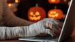 © photo for everything - A mummy's hands typing on a laptop in a spooky, Halloween setting with glowing jack-o'-lanterns in the background. Festive and fun, celebrating Halloween night online.