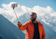 © Anna - Happy Black man taking a selfie with a smartphone in the mountains. Tourist vlogging during an outdoor adventure with a snowy landscape background.