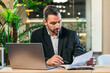 © wedmoments.stock - Businessman in a black suit reviews documents while seated at a desk with a laptop, surrounded by greenery, showcasing a professional work environment and focus on tasks