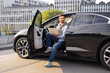 © sofiko14 - A man sits in a black car with a laptop, looking away. The car door is open, and the background includes buildings.