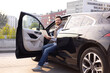 © sofiko14 - A man smiles while working on his laptop inside a black car with the door open, possibly on a rooftop parking area.