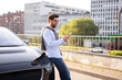 © sofiko14 - A man in glasses leans against a black car, engrossed in his phone, with a city backdrop.