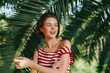 © SHOTPRIME STUDIO - A cheerful young woman stands outdoors among lush palm leaves, wearing a red and white striped top. She smiles warmly, enjoying sunny weather and a carefree summer moment.
