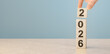 © Celt Studio - Hand building 2026 new year concept with wooden blocks. Male hand placing number two on stack of wood cubes