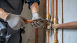 © Anastasiia Trembach - Close-up of a professional plumber repairing copper pipes inside a building, wearing protective gloves and using a pipe wrench to tighten fittings during routine residential plumbing maintenance.