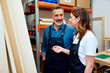 © Seventyfour - Caucasian middle aged man and Caucasian young adult woman standing together in workshop, smiling and talking while working on woodworking project, shelves with materials in background