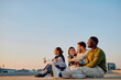 © we.bond.creations - Diverse friends sitting on a rooftop, relaxing and celebrating while drinking wine, enjoying the golden hour cityscape