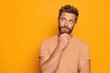 © Darcraft - A bearded man with blue eyes, wearing a striped orange shirt, thoughtfully rests his chin on his hand against a vivid orange background, looking upward with a pondering expression