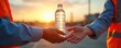 © Viktor - Two hands give water bottle on sunset. Men in safety vests at work. Construction worker offers refreshment after work. Thirsty people drink water. Team delivers liquid. Hydration and energy concept.