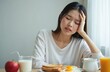 © Viktor - Upset asian girl feels unwell during eating breakfast at home. Woman suffers headache, burnout and depression. Person feel sorrowful and sad, touches head. Mental health and wellness concept.