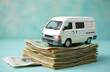 © Viktor - White toy van on top of pile of banknotes on blue background. Stack of paper money. Concept of wealth, finance, savings, transportation, commerce. Toy vehicle on cash. Financial transactions, trade.