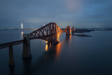 Aerial evening view of the illuminated Forth Bridge in Scotland with reflections on calm water. Iconic landmark captured by drone at dusk.