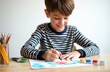 © Maryna - Young boy drawing at desk with colorful paints. Kid draws with focus. Child paints enjoying art. Creative student happy. Education concept. Home schooling activity. Autistic boy draws illustration.