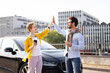 © sofiko14 - A man and woman high-five near a black car, celebrating a moment outdoors in front of a modern building.