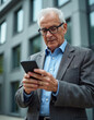 © miss irine - Elegant elderly man uses cellphone on street. Mature senior businessman reads mobile phone screen in city. Modern stylish grandpa types message with smartphone in outdoor urban space.