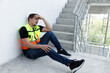 © eakgrungenerd - Construction worker in safety vest sitting on stairway with hands on head, showing signs of stress, fatigue, or headache. Concept of burnout, job pressure, mental health, and labor exhaustion.