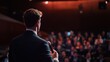 © Bendot - Back view of a speaker holding a microphone, addressing a large, blurred audience in a conference or seminar setting under warm stage lighting.