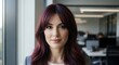 © satrio - Portrait of a confident businesswoman with long, dark red hair standing in a modern office environment.