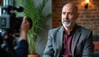 © Maryna - Mature man with grey beard gives interview for camera. Professional filming interview indoors with a cameraperson. Bald man in jacket speaks seriously, looks thoughtful, with plant background.