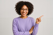 © Marina - Happy and attractive African-American woman in casual lavender sweater and glasses, smiling confidently and pointing finger to the side against a neutral gray background.