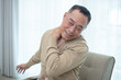 © eakgrungenerd - Senior Asian man sitting on chair with hand on neck, expressing neck pain or muscle stiffness. Concept of elderly health, joint problem or physical discomfort at home.