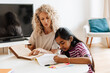 © Drobot Dean - A mother holds a book and watches her daughter write in a notebook while they sit at the table