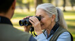 © SYATRI RAWU - A smiling senior woman with grey hair looks through binoculars while on a nature walk, enjoying birdwatching as a hobby with a companion