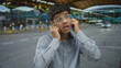 © Krakenimages.com - Young hispanic man wearing glasses and light blue shirt covers ears with hands in crowded modern airport terminal; irritation.