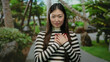 © Krakenimages.com - Woman standing in outdoor park with hands on chest, wearing striped shirt, surrounded by lush greenery, smiling warmly, creating a serene, peaceful atmosphere.