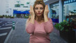 © Krakenimages.com - Woman in pink sweater covering her face with hands and visible fingers while seated before a street building facade; playful shyness.