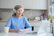 © Liubomir - Senior woman with headphones studies online from her bright home kitchen, smiling and focused while taking notes on a laptop during a webinar or virtual class, lifelong learning
