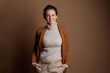 © Alliance - A smiling businesswoman in her 30s-40s, dressed in a brown blazer and beige trousers, stands with hands in pockets, exuding a confident and approachable demeanor in this studio portrait.