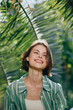 © SHOTPRIME STUDIO - Smiling woman wearing a green striped jacket stands among lush tropical plants under bright sunlight, creating a fresh, summery portrait.