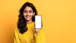 © dana - A smiling Indian woman in a sweater holds a smartphone with a blank screen against a yellow background. A beautiful young woman shows a phone with a white screen
