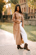 © Darius - Stylish woman in a brown coat walks on a sunny street in autumn, creating a chic urban vibe