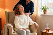 © Ira - Serene mature woman sits by window as partner gently massages her shoulders, creating atmosphere of warmth, care, and comfort during menopause phase at home.