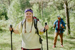 © AnnaStills - Portrait of young Caucasian girl with Down syndrome hiking in forest, holding trekking poles and wearing backpack, young adult man with beard walking behind her in background