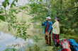 © AnnaStills - Caucasian young adult man and Caucasian young girl with Down syndrome standing on grassy riverbank fishing together, holding fishing rod, surrounded by trees and outdoor gear, looking at water