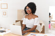 © etonastenka - African American woman sitting at a desk in a bright modern apartment, working on a laptop and smiling at the camera. Concept of remote work, communication, technology, and positive online interaction