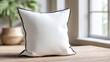 © Mommangophoto - A cozy white pillow with a black trim sits on a wooden table, surrounded by natural light and greenery in a serene indoor setting.