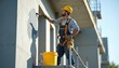 © Vadym - Craftsman applies grey paint to exterior wall on build. Hardhat worker stands on scaffold. Man repairs concrete wall, wears safety harness on construction site. Uses brush for facade repair. Builder