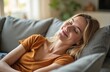 © Vadym - Woman lies on gray couch, head on pillow, eyes closed. Wears orange t-shirt. Blonde hair. Woman smiles, looks relaxed, peaceful. Calm expression. Sunny home interior with window, plant in background.