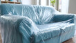 © sornram - A new blue sofa covered in plastic wrap, waiting to be unveiled in a cozy living room with natural light.