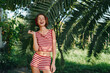 © SHOTPRIME STUDIO - A cheerful woman wearing a striped red and white two piece stands beneath lush palm fronds in a sunny outdoor setting, smiling playfully and posing with carefree confidence.